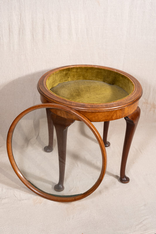 An antique round wooden side table with three cabriole legs, featuring a gold-leafed velvet upholstered top and a matching round wooden mirror leaning against it, set against a draped beige fabric backdrop.