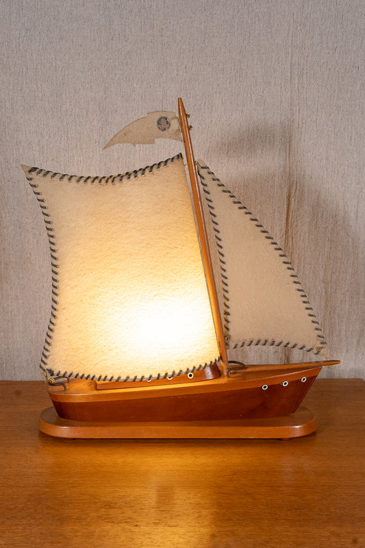 A wooden model sailboat lamp with illuminated cream-colored sails, displayed on a wooden surface against a neutral textured wall.