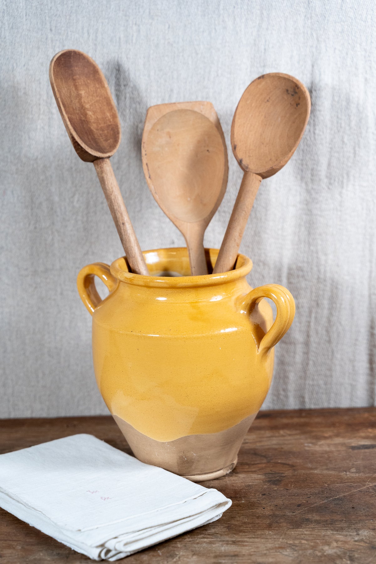 Three wooden kitchen utensils, including two spoons and a spatula, arranged in a glossy yellow ceramic pot with side handles, set on a rustic wooden table with a folded white cloth in front.