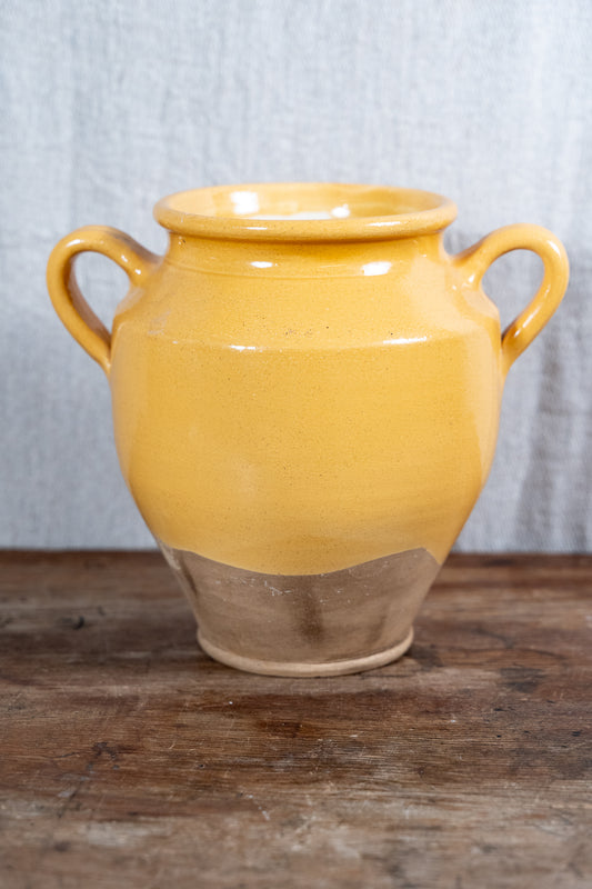 A handmade ceramic vase with a glossy mustard yellow glaze and a natural unglazed clay base, featuring two side handles, set on a rustic wooden surface against a gray textured backdrop.
