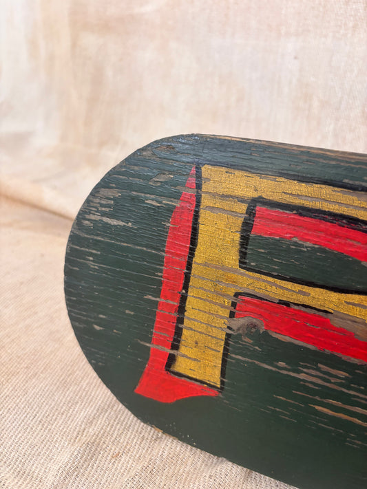 A close-up of a dark green wooden sign featuring a large distressed yellow and red painted letter 'F' on a beige fabric background.