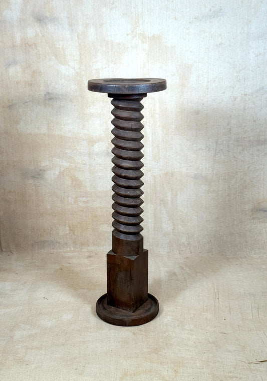 A rustic dark wood pedestal stand shaped like a giant threaded screw, featuring a circular top and hexagonal base, set against a textured beige backdrop.