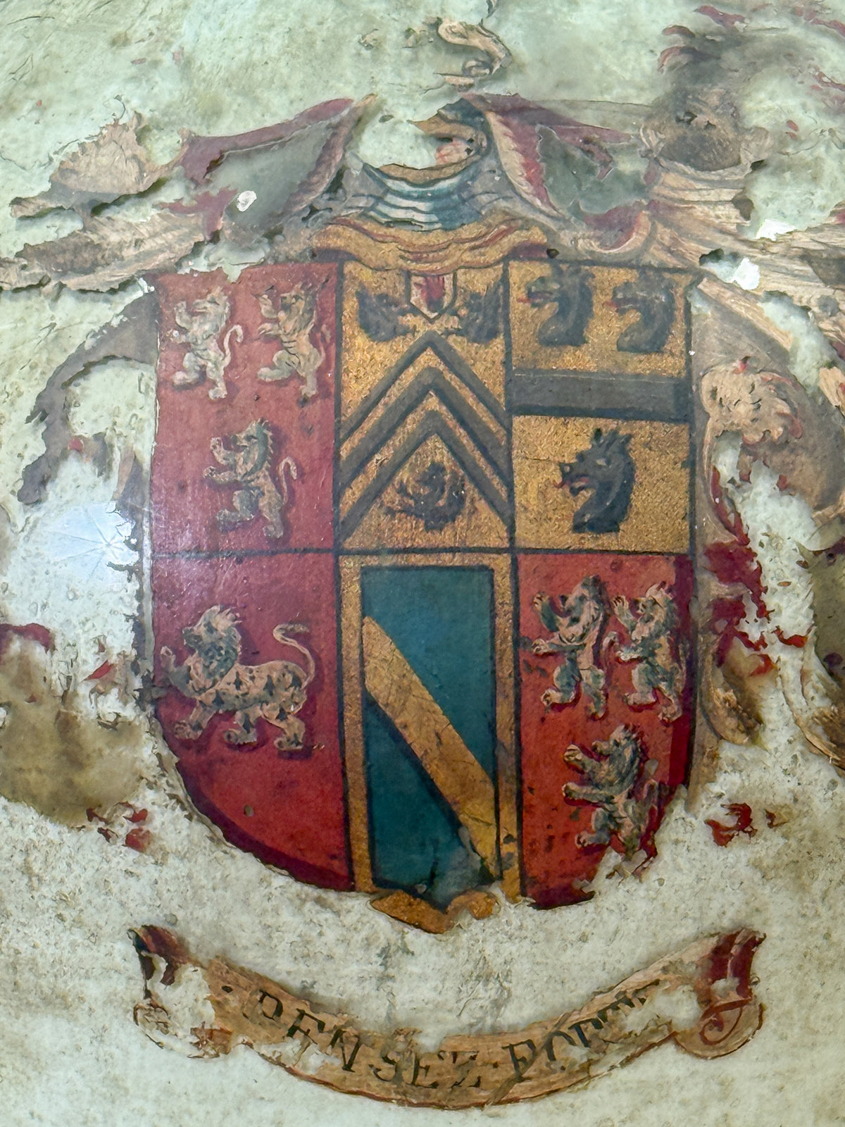 An antique painted heraldic crest on a weathered glass surface, featuring red, gold, black, and teal sections with lions, chevrons, and a banner below.