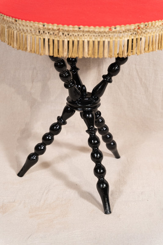 A close-up of a round table with a bright red upholstered top and gold fringe trim, supported by a glossy black turned spindle-leg base on a light fabric backdrop.