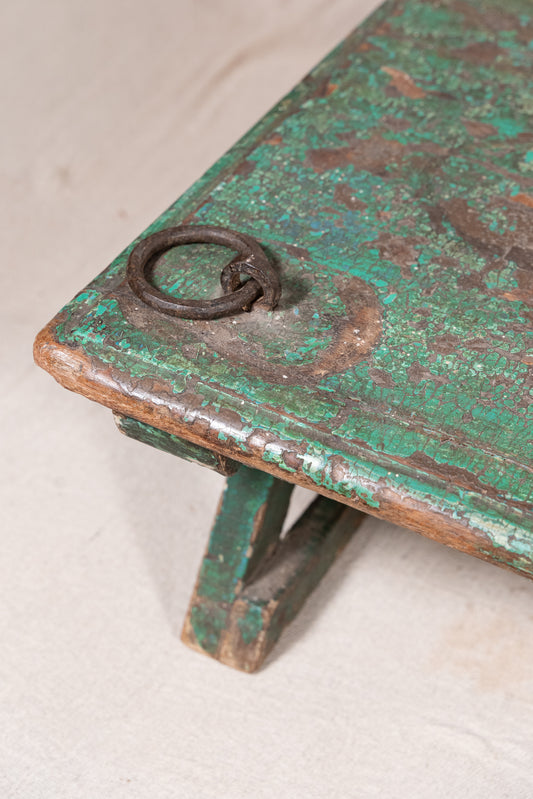 Low wooden bajot table with distressed green paint, angled legs and metal corner rings, possibly repurposed from an old gate or door.