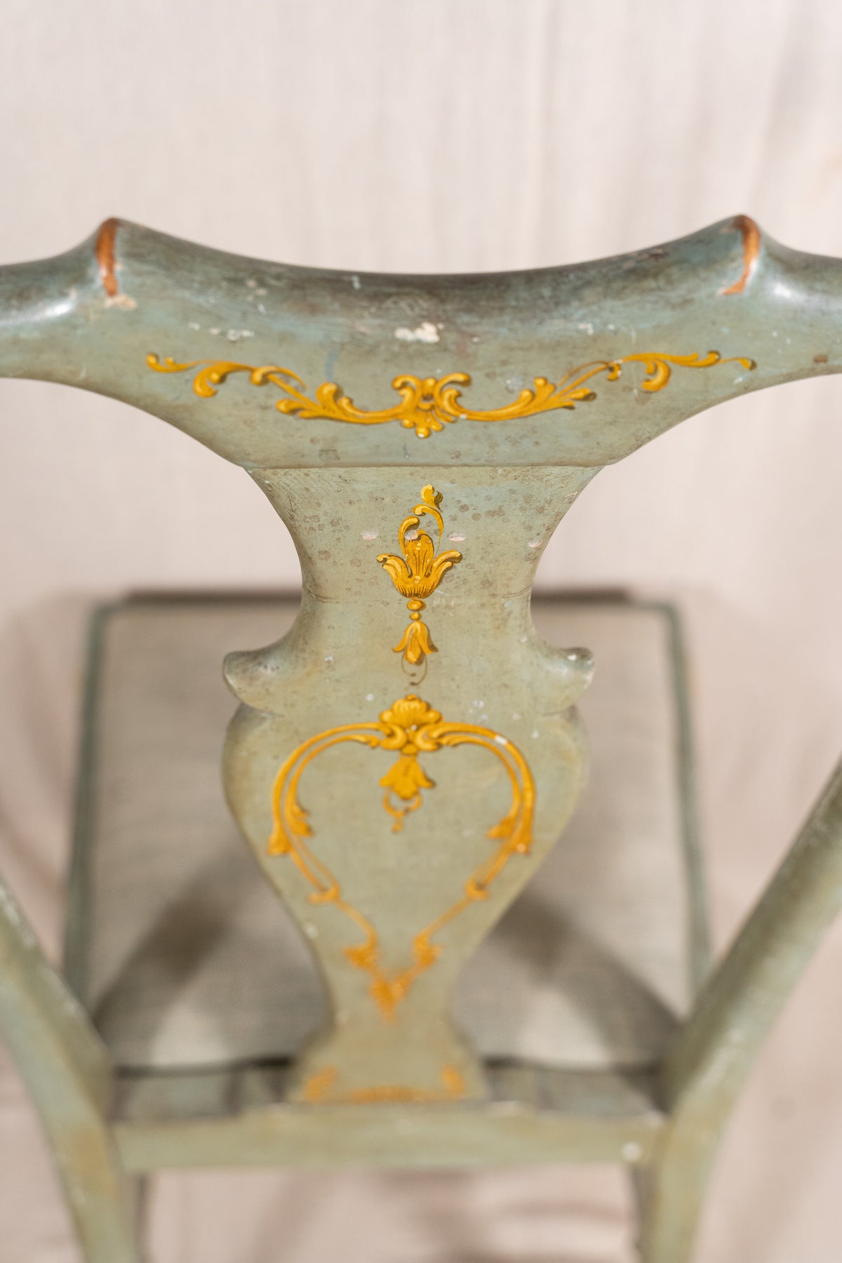 A close-up of a vintage wooden chair back painted in a distressed sage green with ornate gold floral scrollwork details.