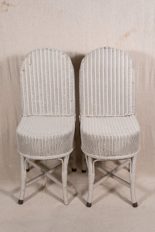 A pair of white wicker dining chairs with high arched backs and crossed leg frames, set against a neutral beige fabric backdrop.