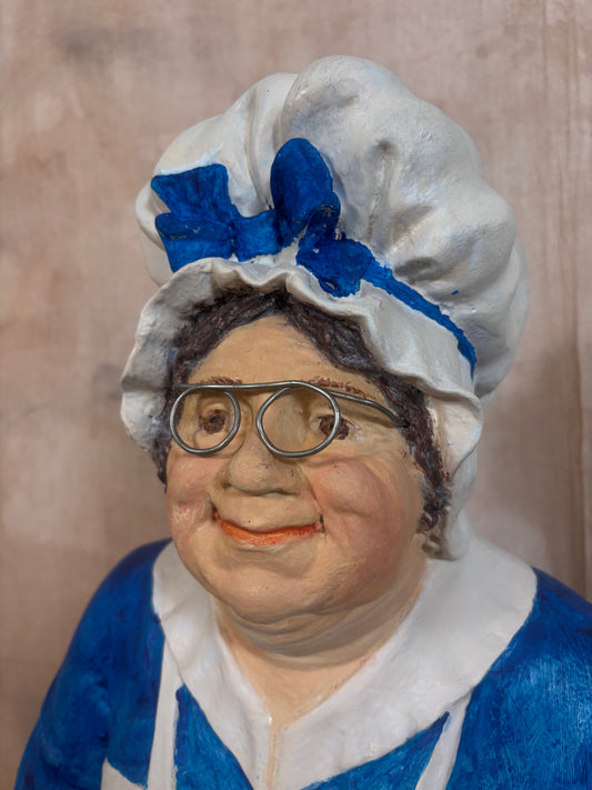 A close-up portrait of a decorative figurine or sculpture depicting an older woman with round glasses, a white ruffled bonnet with a blue bow, and a blue-and-white dress, set against a textured beige background.