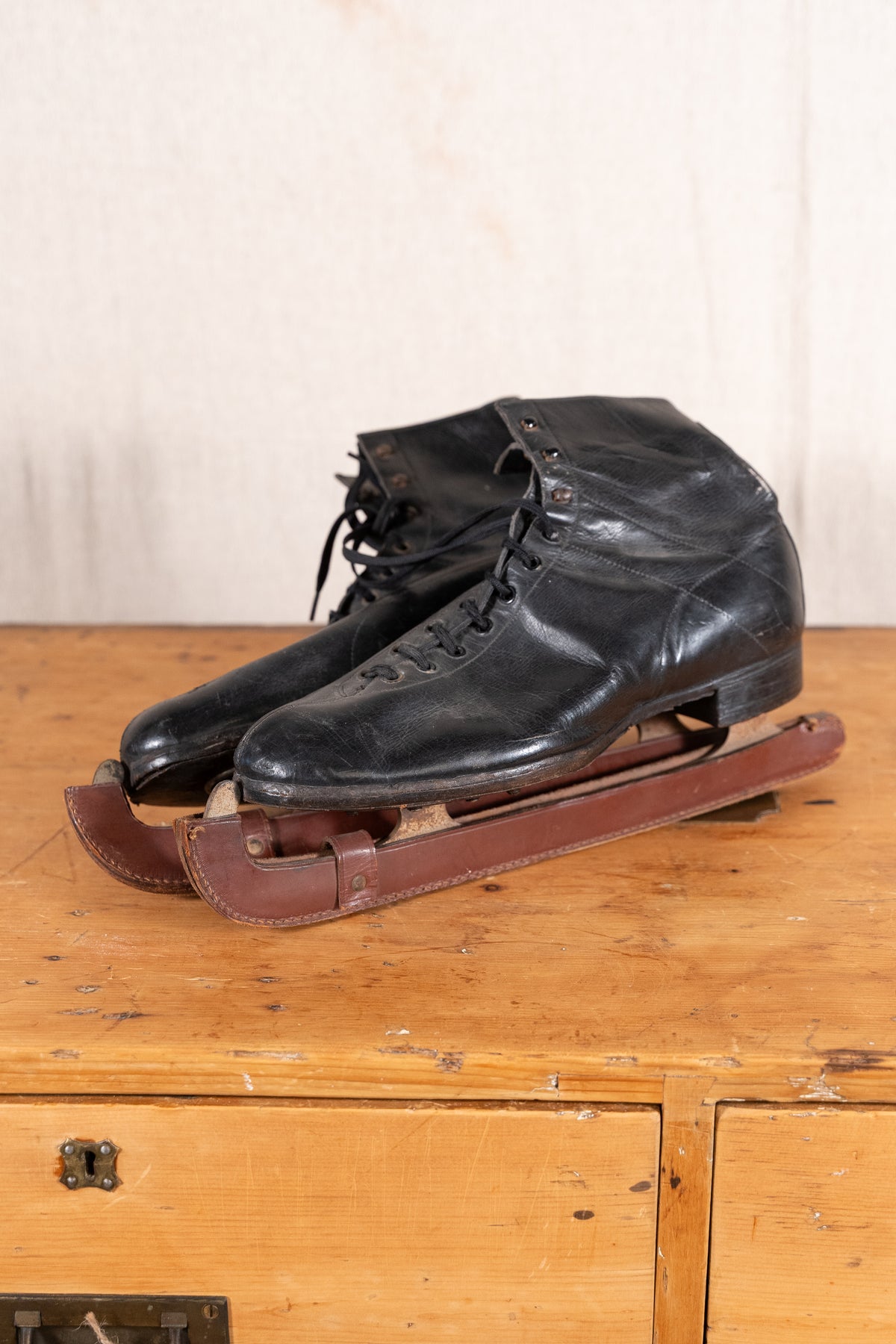 A pair of vintage black leather ice skates with brown wooden blades, displayed on a rustic wooden chest against a light beige wall.