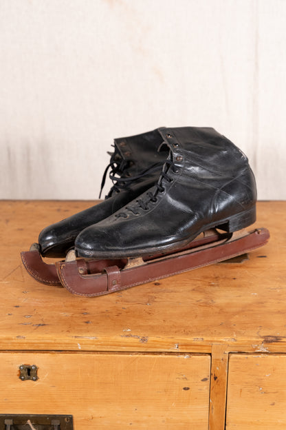 A pair of vintage black leather ice skates with brown wooden blades, displayed on a rustic wooden chest against a light beige wall.