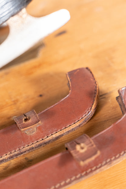 Close-up of two brown leather straps with stitched edges and small metal rivets, laid on a wooden surface.