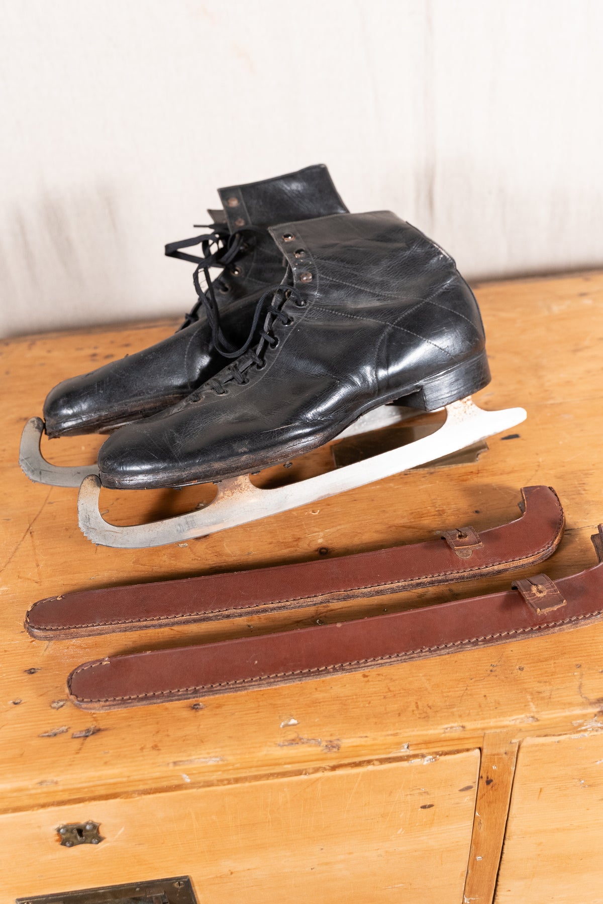 A pair of vintage black leather ice skates with metal blades, displayed on a wooden surface with brown leather skates or blade covers placed in front.