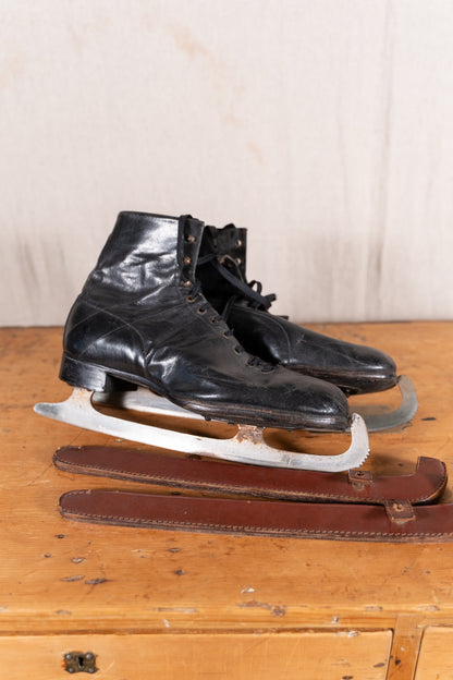 A pair of vintage black leather ice skates with metal blades, resting on a wooden surface with a brown leather strap in front of them.