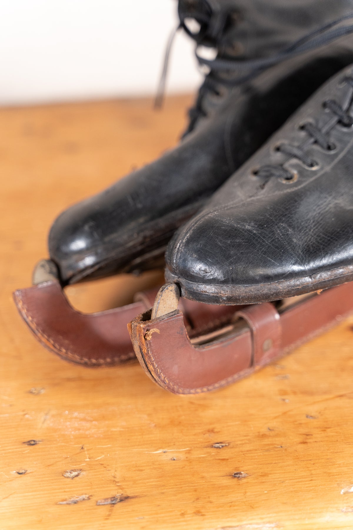 A close-up of a pair of vintage black leather ice skates with brown leather blades resting on a wooden surface.