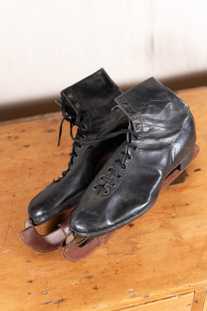 A pair of vintage black leather ice skates with brown leather straps and metal blades, resting on a rustic wooden table.