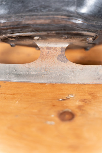 Close-up of a vintage metal skate blade resting on a wooden surface, with part of a dark boot or skate component visible at the top.