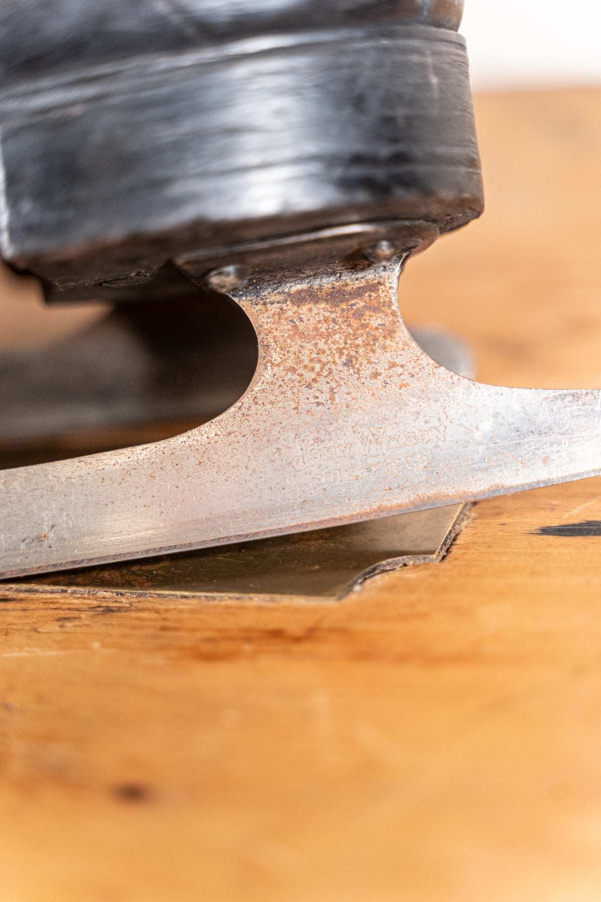A close-up shot of a vintage metal ice skate blade resting on a worn wooden surface, showing the aged, rusted metal and dark rubber boot.