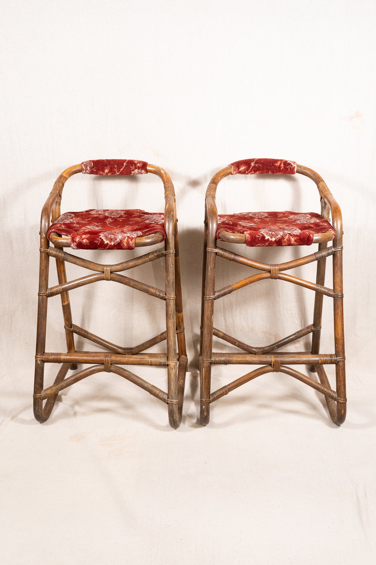 Pair of vintage bamboo bar stools with dark finished frames, red patterned velvet-style upholstery, X-shaped cross supports and lower footrests.