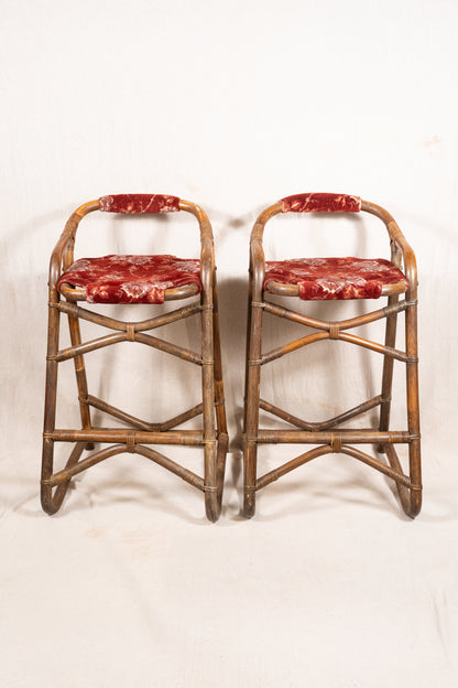 Pair of vintage bamboo bar stools with dark finished frames, red patterned velvet-style upholstery, X-shaped cross supports and lower footrests.