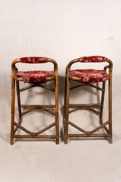 Pair of vintage bamboo bar stools with dark finished frames, red patterned velvet-style upholstery, X-shaped cross supports and lower footrests.