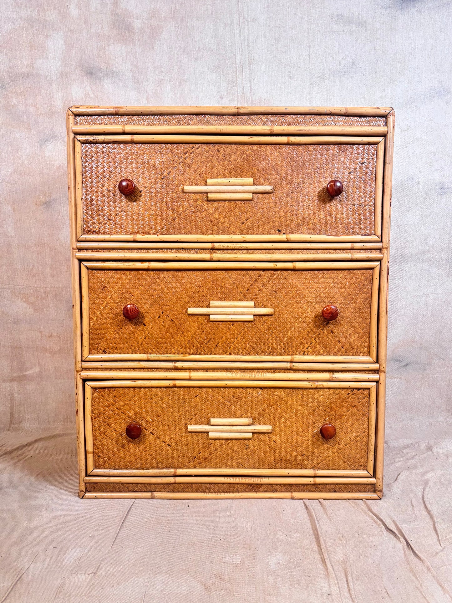 A three-drawer rattan chest of drawers with a natural bamboo frame and reddish-brown round knobs, set against a neutral fabric backdrop.