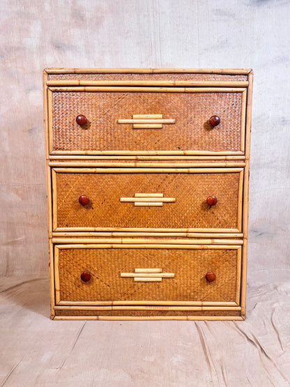 A three-drawer rattan chest of drawers with a natural bamboo frame and reddish-brown round knobs, set against a neutral fabric backdrop.