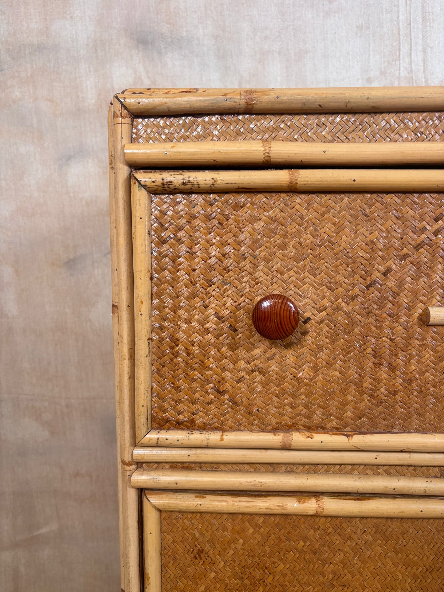 Close-up of a rattan and bamboo chest or cabinet drawer, showing woven natural-fiber panels, a round wooden knob, and light bamboo framing against a neutral textured wall.