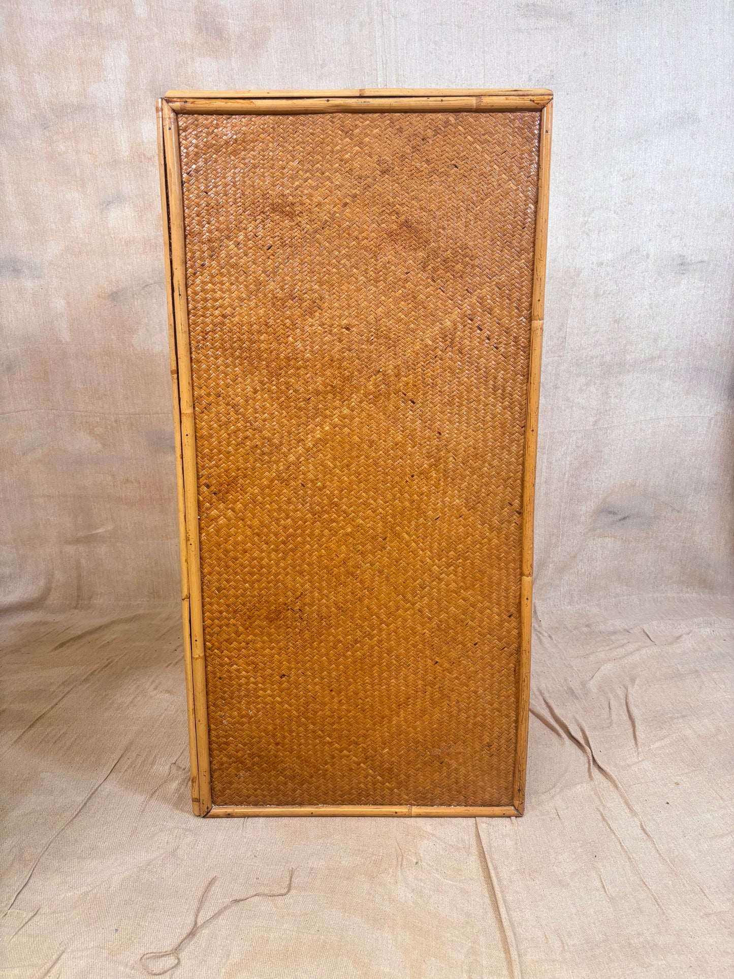 A rectangular vintage rattan or woven cane panel with a natural bamboo frame, standing upright against a neutral beige fabric backdrop.