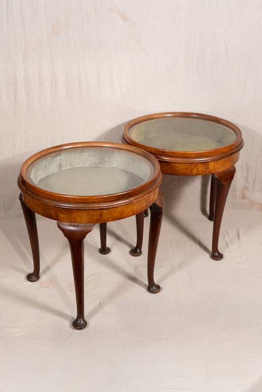 A pair of vintage wooden side tables with oval glass-top surfaces and curved cabriole-style legs, photographed against a neutral draped fabric backdrop.