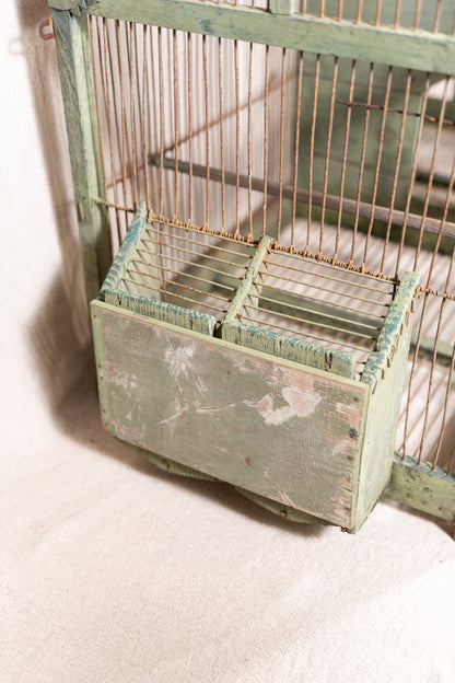 Vintage wooden birdcage with weathered green paint, decorative metal ridge detail, wire mesh panels and feeding troughs on both sides.