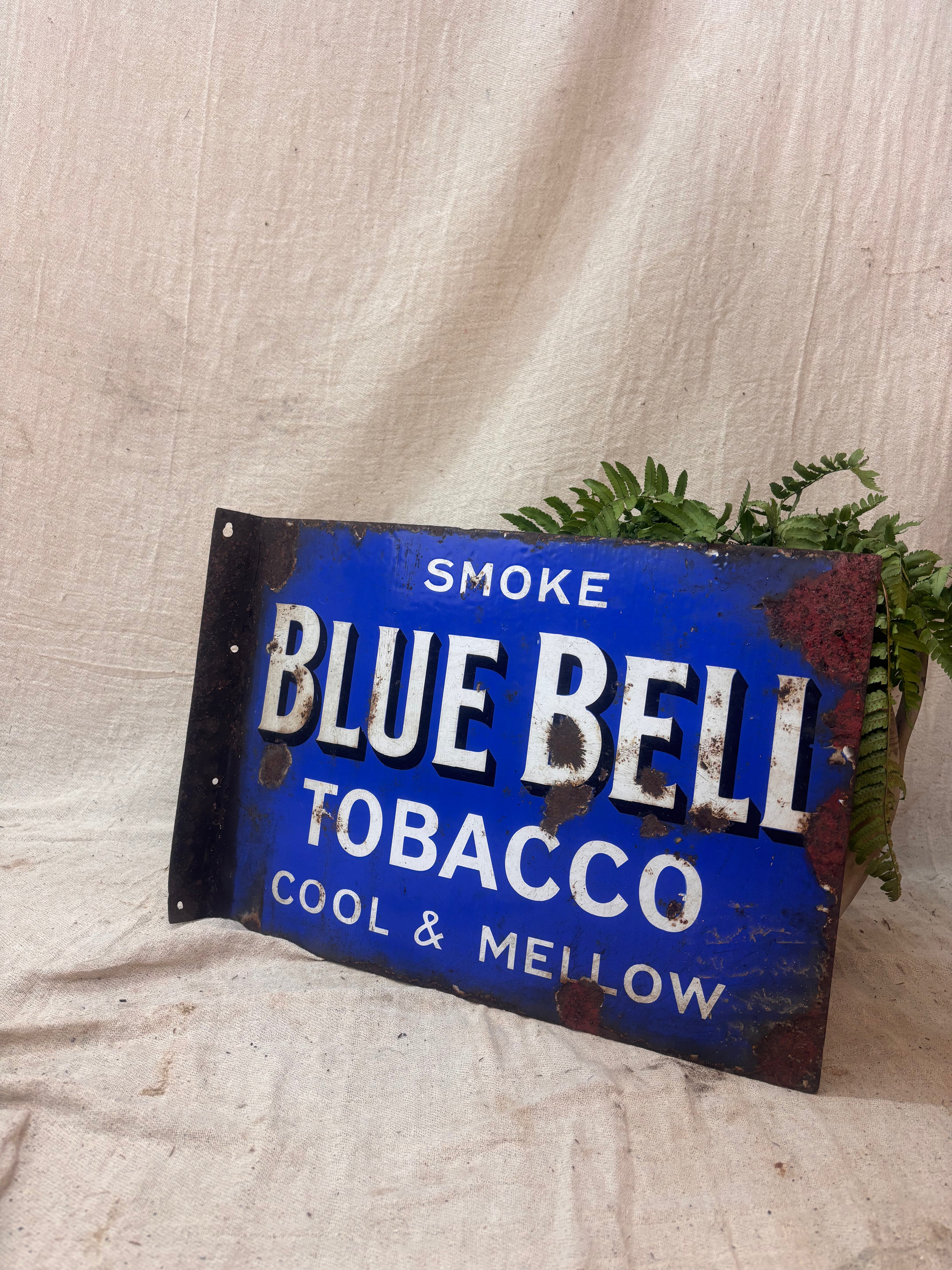 Vintage Blue Bell Tobacco sign with text 'Smoke Blue Bell Tobacco Cool & Mellow' on a textured fabric background.