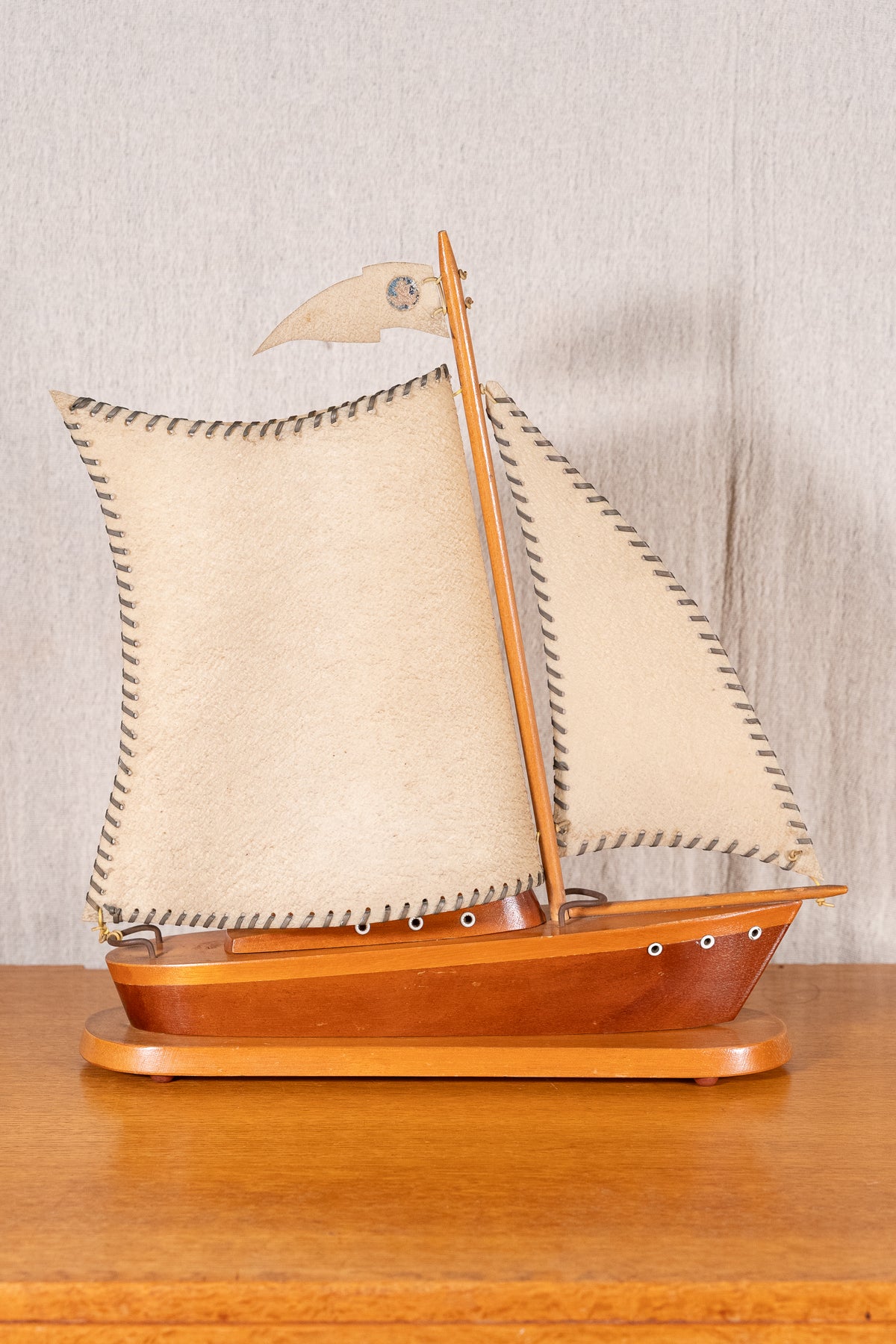 A wooden model of a traditional sailing ship with beige canvas sails and a polished reddish-brown hull, displayed on a wooden surface against a neutral textured wall.