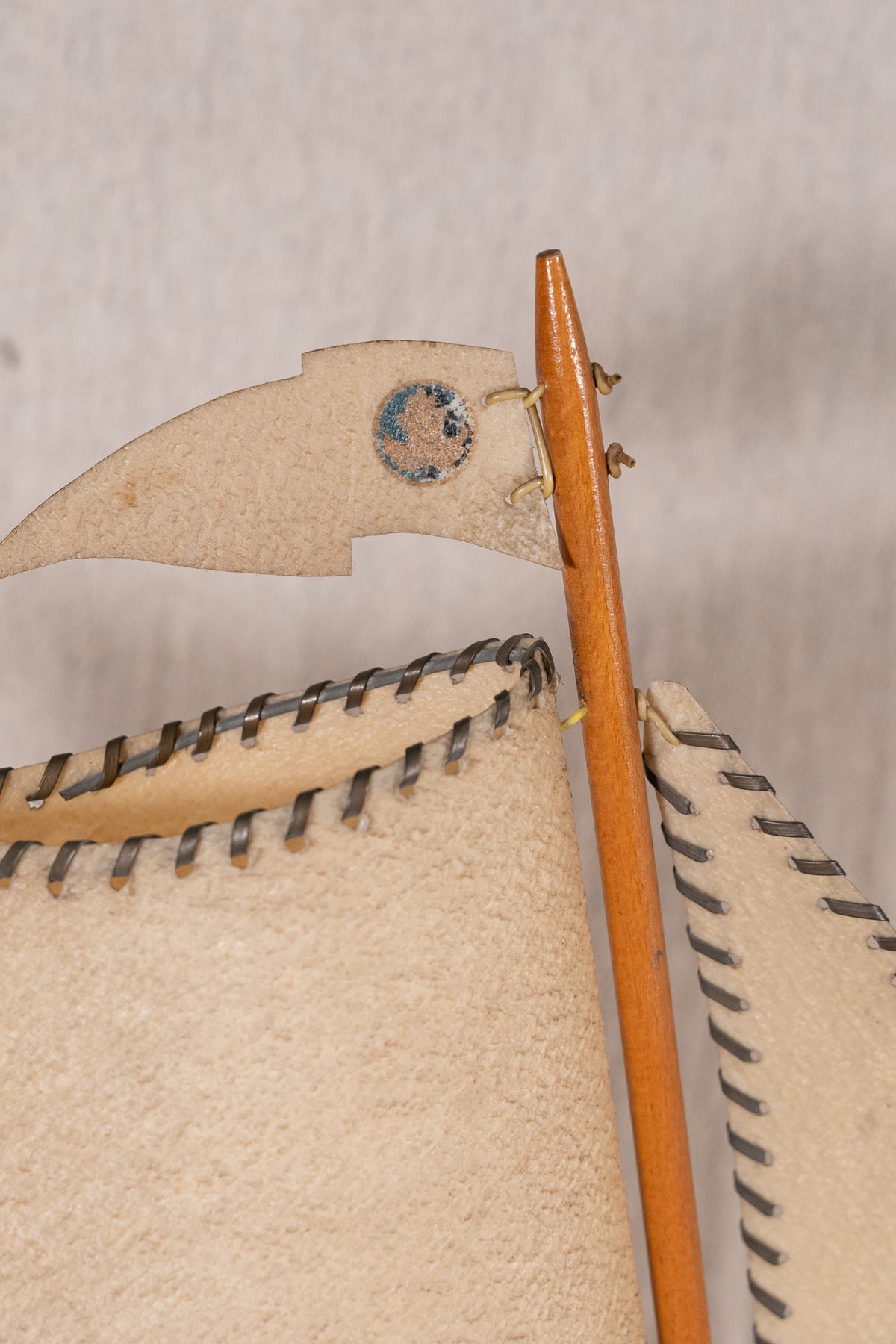 Close-up of a handmade wooden toy or model ship with beige stitched sails and a light brown wooden mast against a neutral light gray background.