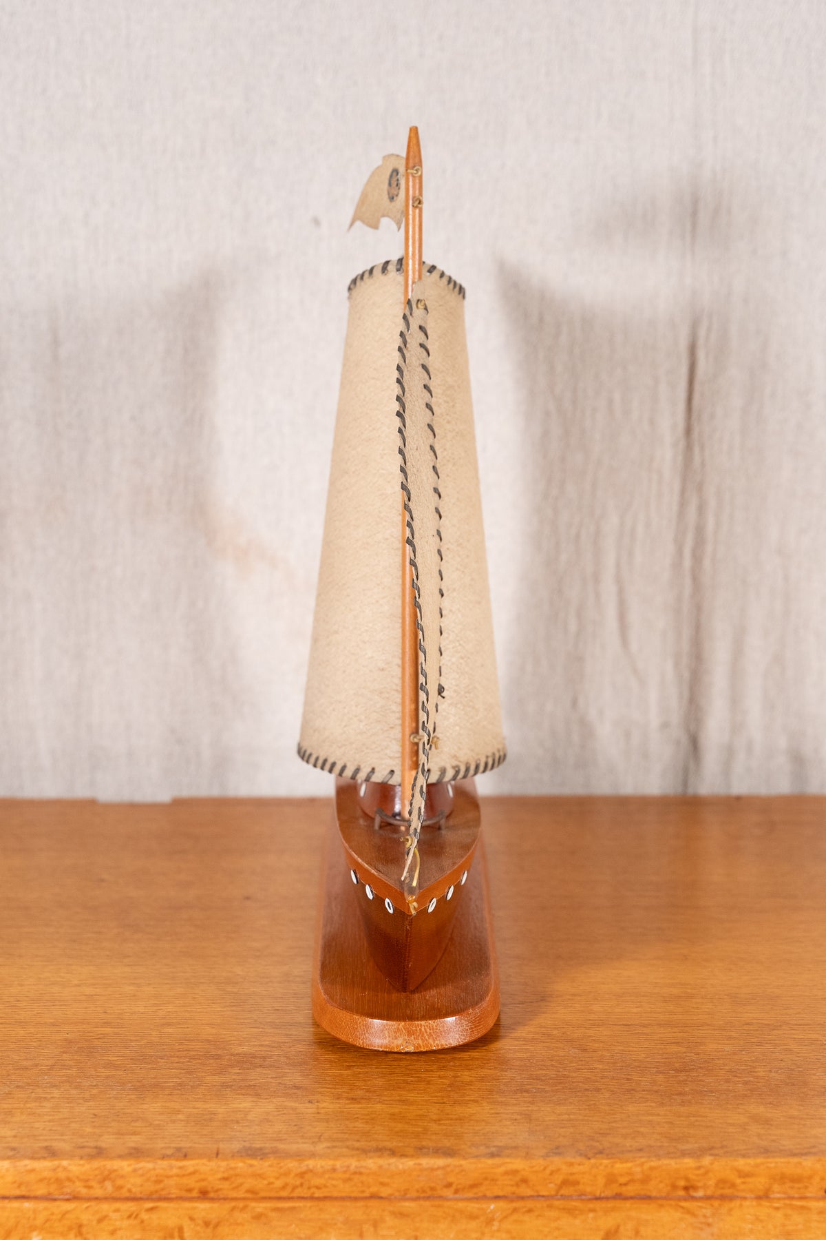 A miniature wooden model sailboat with a beige canvas sail and a polished brown wooden hull, displayed on a wooden surface against a neutral fabric backdrop.