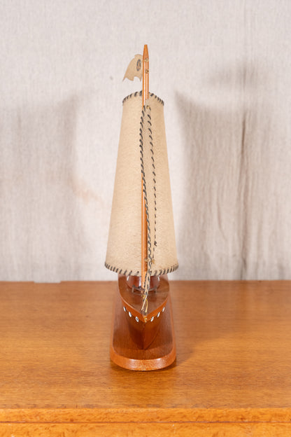 A miniature wooden model sailboat with a beige canvas sail and a polished brown wooden hull, displayed on a wooden surface against a neutral fabric backdrop.