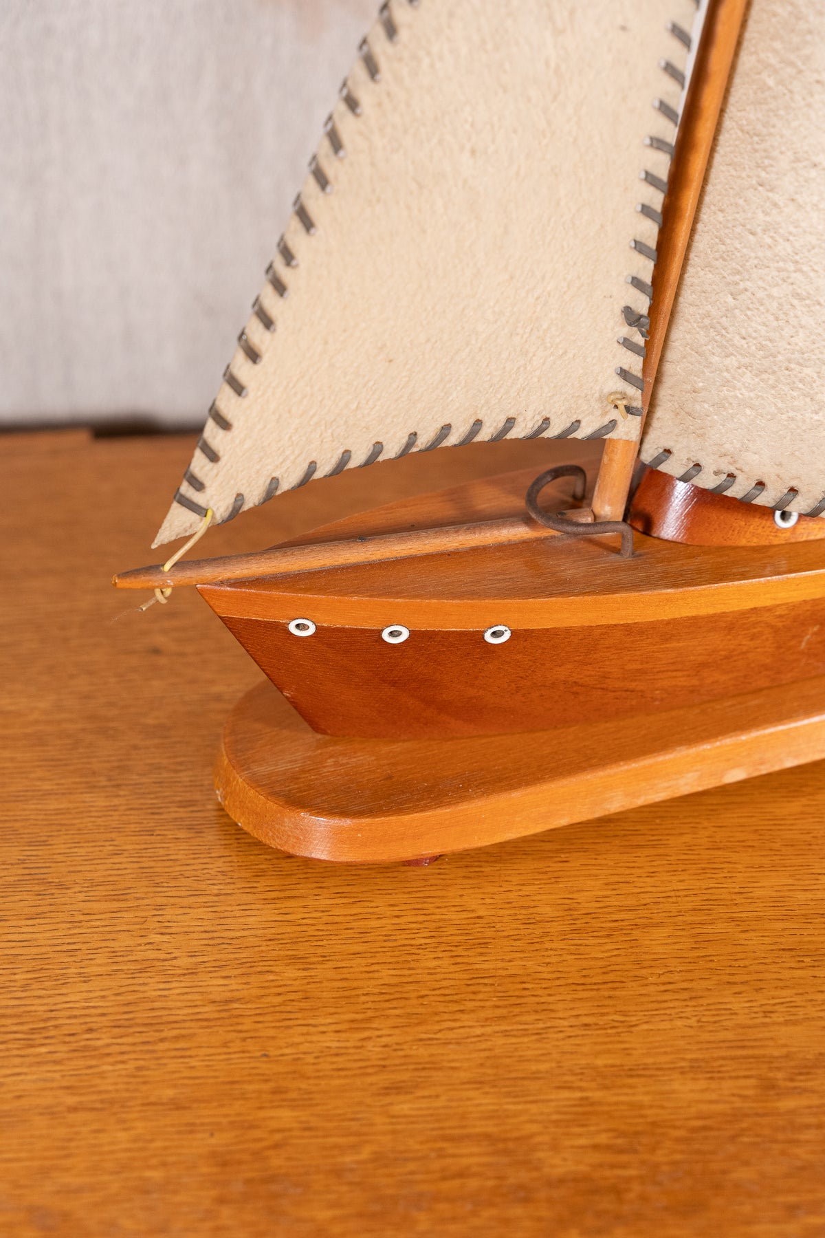 A close-up of a wooden model sailboat with cream-colored sails, resting on a wooden tabletop.