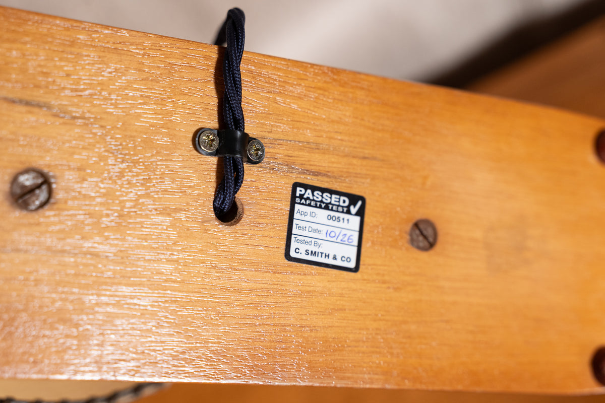 Close-up of a wooden boat deck with a black rope tied to a metal cleat and a small black 'PASSED V' warranty label attached to the wood grain surface.