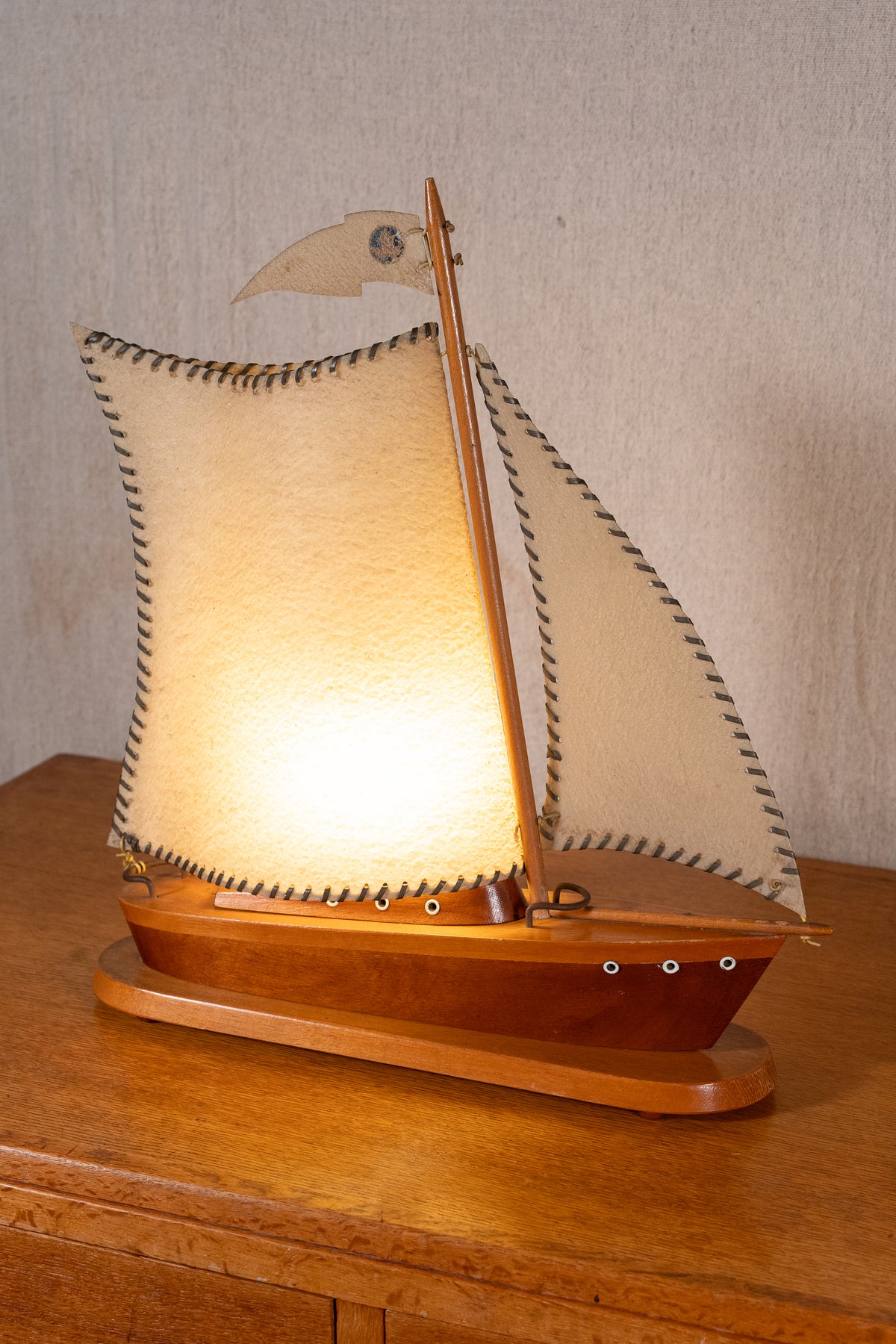 A wooden model sailboat with illuminated cream-colored fabric sails, displayed on a wooden surface against a neutral textured wall.