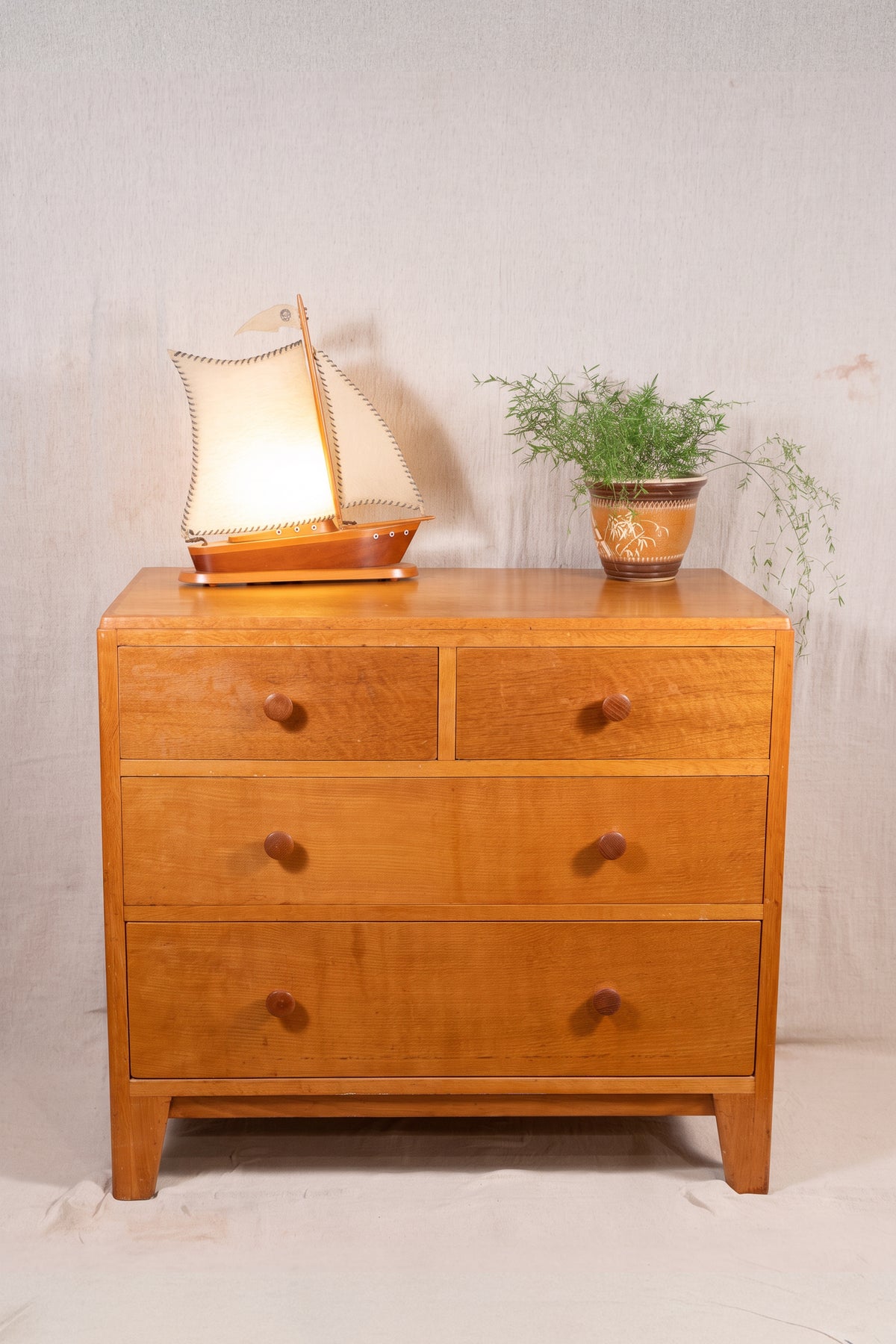 A mid-century modern wooden chest of drawers with three drawers, styled with a glowing model sailboat and a potted plant on top, set against a neutral light gray backdrop.