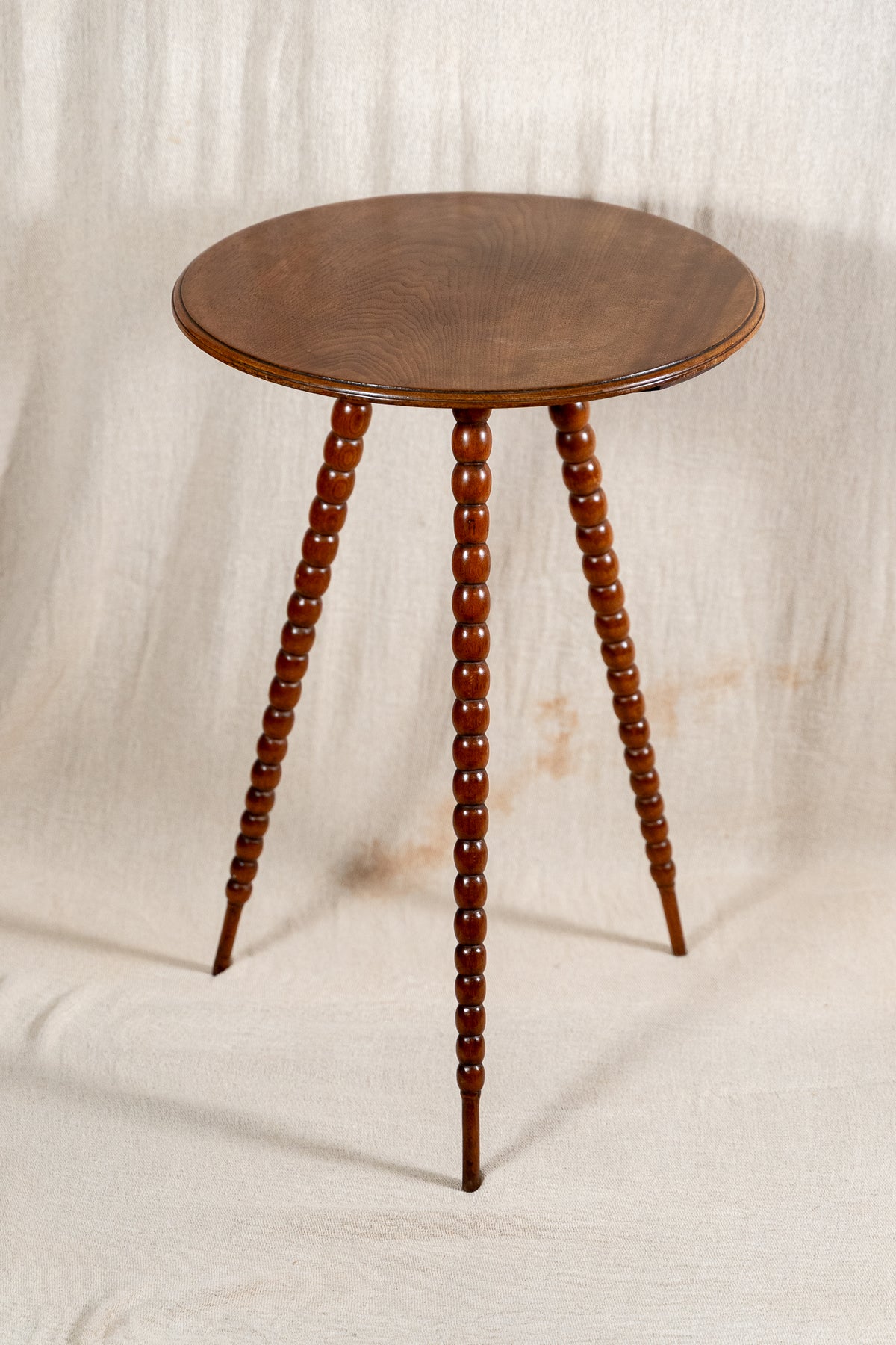 A round wooden side table with three turned spindle legs, shown against a draped beige fabric backdrop.