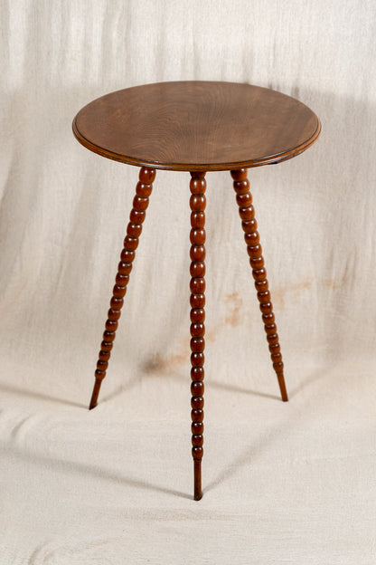 A round wooden side table with three turned spindle legs, shown against a draped beige fabric backdrop.