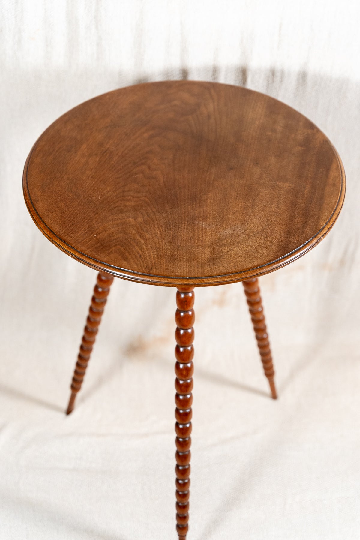A round wooden side table with a medium brown finish and three turned spindle legs, photographed against a draped white fabric backdrop.