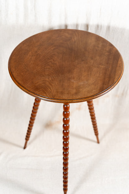 A round wooden side table with a medium brown finish and three turned spindle legs, photographed against a draped white fabric backdrop.