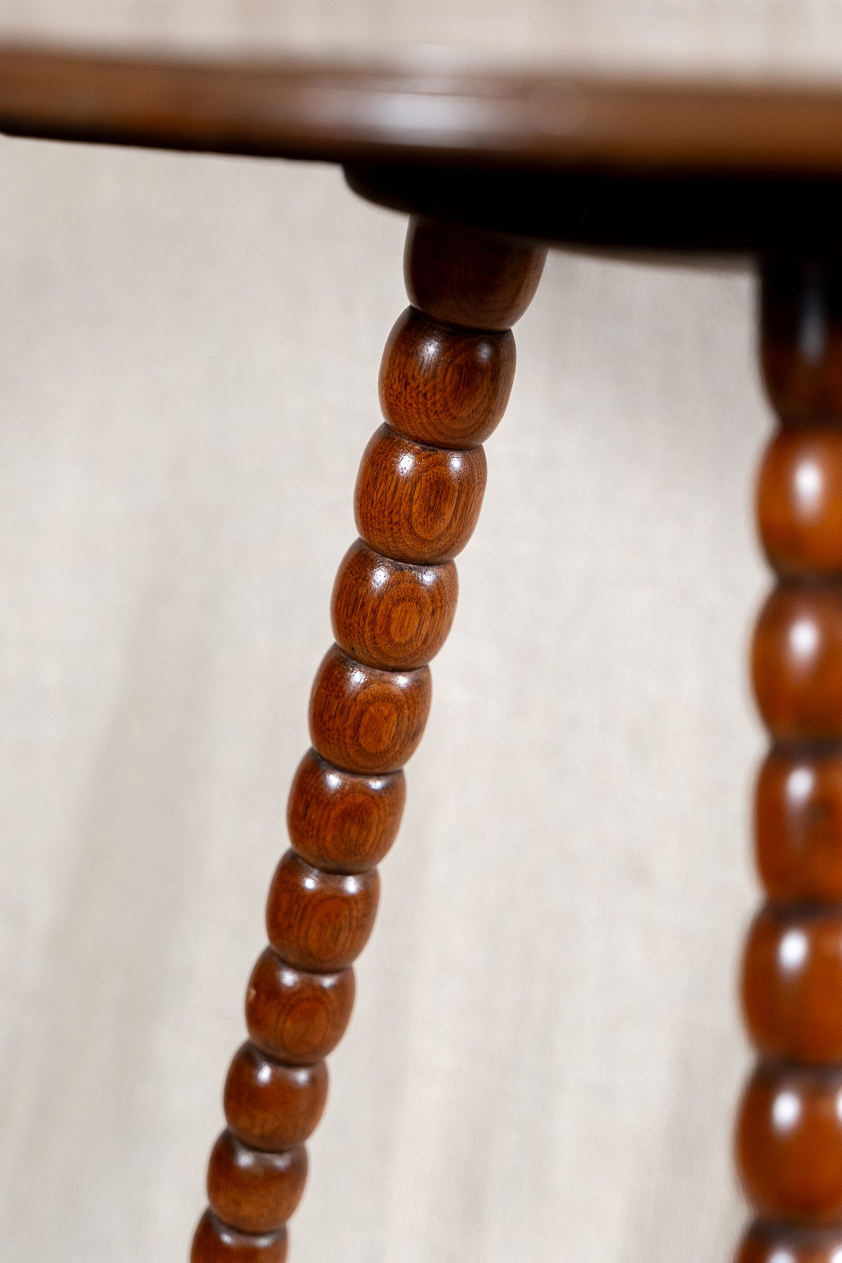 Close-up of a dark reddish-brown wooden table leg with a series of turned oval beads, set against a neutral beige fabric background.