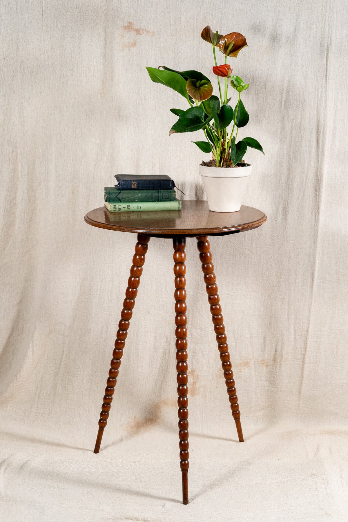 A small round wooden side table with three turned spindle legs, styled with a potted anthurium plant and a stack of books, set against a draped beige fabric backdrop.
