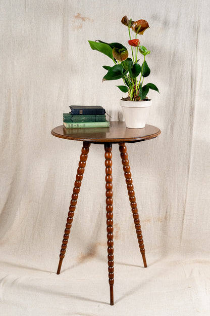 A small round wooden side table with three turned spindle legs, styled with a potted anthurium plant and a stack of books, set against a draped beige fabric backdrop.
