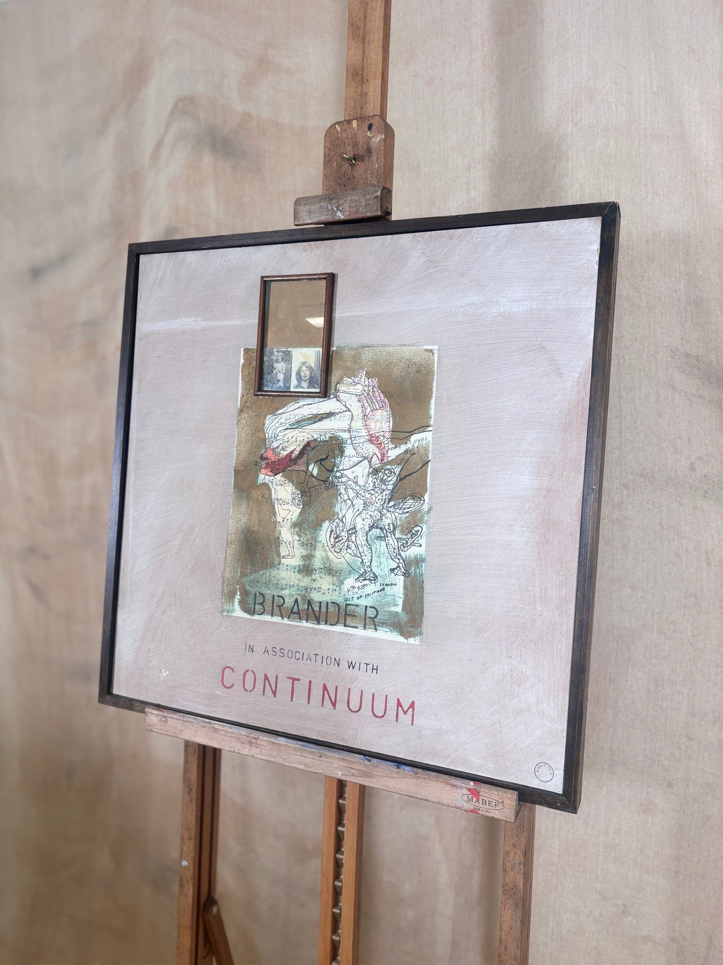 A framed art print or mixed-media piece displayed on a wooden easel against a light wood wall. The artwork features a textured beige background, a central collage-like illustration with a small mirror inset, and the text 'BRANDER IN ASSOCIATION WITH CONTINUUM' in red and black.