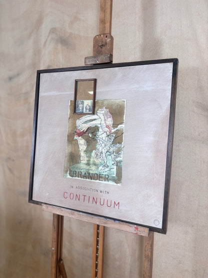 A framed art print or mixed-media piece displayed on a wooden easel against a light wood wall. The artwork features a textured beige background, a central collage-like illustration with a small mirror inset, and the text 'BRANDER IN ASSOCIATION WITH CONTINUUM' in red and black.