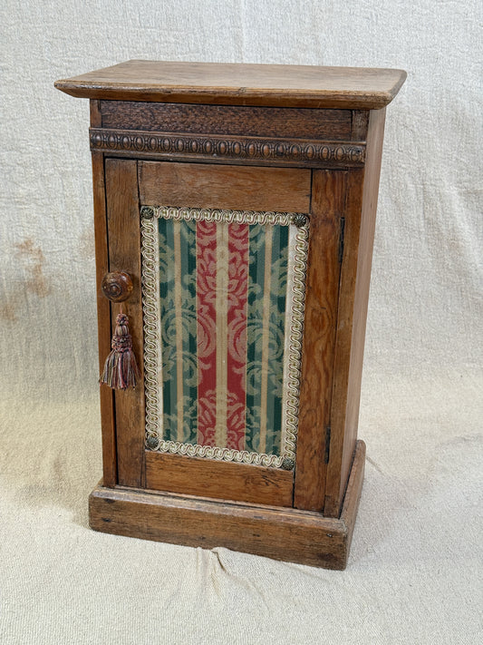 Wooden bedside cabinet with patterned fabric-inset door, braided gimp trim, tassel knob and carved egg-and-dart detailing. 
