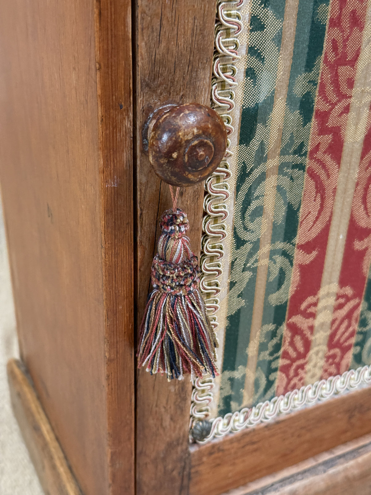 Wooden bedside cabinet with patterned fabric-inset door, braided gimp trim, tassel knob and carved egg-and-dart detailing. 